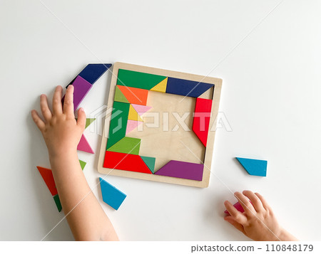 childs hand collects multicolored wooden mosaic on white background. child solves colorful tangram childs hand collects multicolored wooden mosaic on white background. child solves colorful tangram 110848179