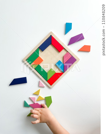 childs hand collects multicolored wooden mosaic on white background. child solves colorful tangram 110848209