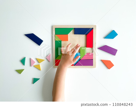 childs hand collects multicolored wooden mosaic on white background. child solves colorful tangram childs hand collects multicolored wooden mosaic on white background. child solves colorful tangram 110848243