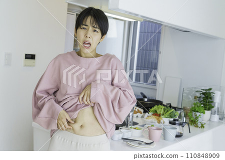 A young woman pinching her stomach in front of the kitchen and worrying about fat 110848909