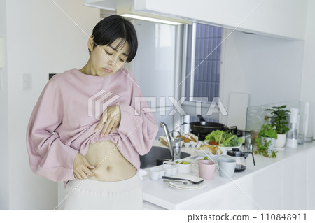 一位年輕女子在廚房前捏著肚子，擔心脂肪 110848911