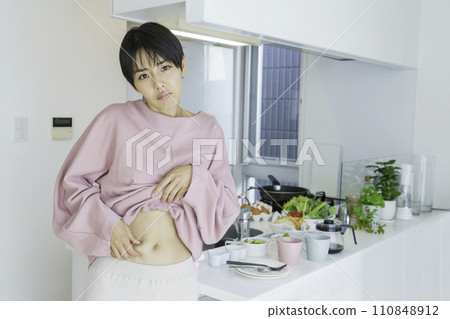一位年輕女子在廚房前捏著肚子，擔心脂肪 110848912