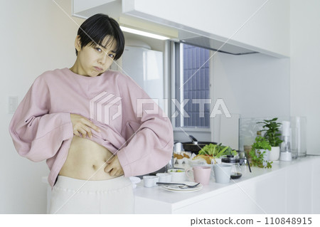 一位年輕女子在廚房前捏著肚子，擔心脂肪 110848915