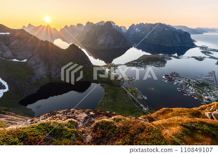 Sunset casts radiant light over Reinebringen, with the sun mirroring on calm fjord waters 110849107