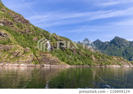A waterfall cascades in Trollfjorden, flanked by rugged mountains and mirror-like waters A waterfall cascades in Trollfjorden, flanked by rugged mountains and mirror-like waters 110849124