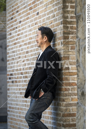 A man in his mid-50s wearing a black jacket leaning against a brick wall. A man in his mid-50s wearing a black jacket leaning against a brick wall. 110849800