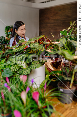 Vertical attractive and smiling young asian busy businesswoman, owner of flower shop. Spraying water and fertilizing herbs in pots. Vertical attractive and smiling young asian busy businesswoman, owner of flower shop. Spraying water and fertilizing herbs in pots. 110850398