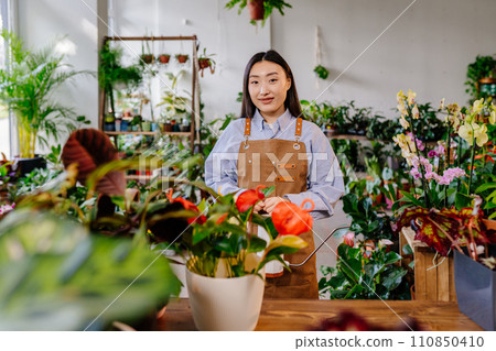 Startup small business concept. Portrait of young korean woman owner standing wearing apron behind counter in flower plant shop interior. Startup small business concept. Portrait of young korean woman owner standing wearing apron behind counter in flower plant shop interior. 110850410
