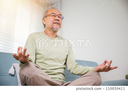 Lifestyle senior man sitting on sofa in living room holding hands in mudra practicing home yoga in lotus pose, Elderly relaxed man do physical exercises following healthy on stress free weekend Lifestyle senior man sitting on sofa in living room holding hands in mudra practicing home yoga in lotus pose, Elderly relaxed man do physical exercises following healthy on stress free weekend 110850411