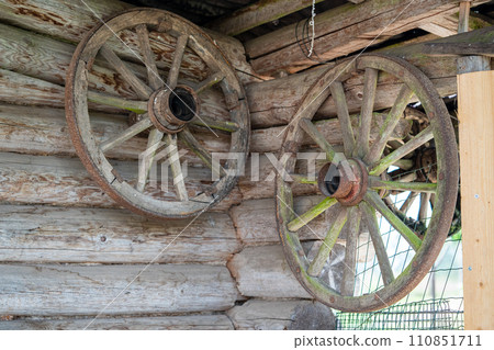 An old wooden cart wheels hang on the log wall of a village house. An old wooden cart wheels hang on the log wall of a village house. 110851711