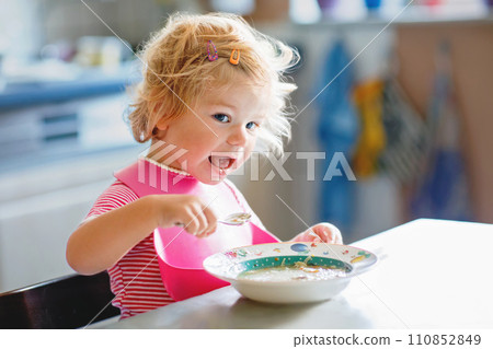 Adorable baby girl eating from spoon vegetable noodle soup. Healthy food, child, feeding and development concept. Cute toddler child with spoon sitting in highchair and learning to eat by itself 110852849