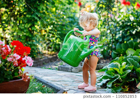 Cute little baby girl in colorful swimsuit watering plants and blossoming flowers in domestic garden on hot summer day. Adorable toddler child having fun with playing with water and ca 110852853