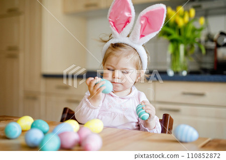 Cute little toddler girl wearing Easter bunny ears playing with colored pastel eggs. Happy baby child unpacking gifts. Adorable healthy smiling kid in pink clothes enjoying family holiday Cute little toddler girl wearing Easter bunny ears playing with colored pastel eggs. Happy baby child unpacking gifts. Adorable healthy smiling kid in pink clothes enjoying family holiday 110852872