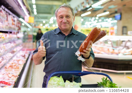 Mature man diligently choosing tasty sausage in supermarket Mature man diligently choosing tasty sausage in supermarket 110854428