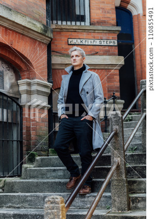 Handsome male model with gray hair in a black roll neck jacket and trousers, checked coat walking on the old city street. Cold season fashion stylish outfit trend. Old money aesthetic. Soft focus Handsome male model with gray hair in a black roll neck jacket and trousers, checked coat walking on the old city street. Cold season fashion stylish outfit trend. Old money aesthetic. Soft focus 110855491