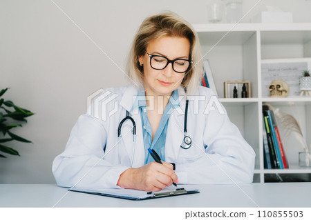 Confident mature middle aged woman doctor medical nurse, female physician practitioner in white coat with stethoscope sitting at working place at hospital office and making notes during appointment. 110855503