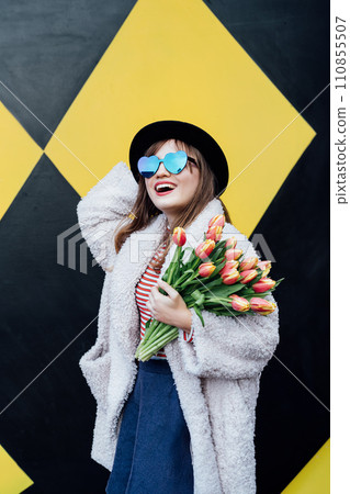 Happy emotional young fashion woman in heart-shaped sunglasses holding bouquet of fresh tulip flowers on the geometry bright yellow and black wall background. Urban city street fashion. Spring mood. 110855507