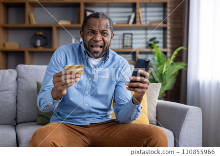 African american man sitting on sofa while holding mobile phone and plastic bank card with stressed facial expression. Adult male buyer making unsuccessful attempts paying for online order at home. 110855966