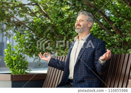 A poised senior businessman in a suit takes a tranquil break outdoors, practicing mindfulness amidst the city setting. 110855986