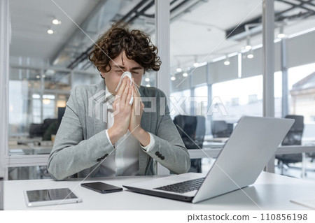 Exhausted young adult businessman caught a cold, working with a laptop at the office and sneezing into a tissue. Displaying common office work fatigue. Exhausted young adult businessman caught a cold, working with a laptop at the office and sneezing into a tissue. Displaying common office work fatigue. 110856198