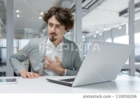 Professional young adult male office worker looking uneasy or stressed, with his hand on the chest, possibly experiencing burnout or health issues at work. 110856206