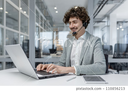 Smiling young businessman with headset working on laptop in modern office, showcasing multitasking and customer service skills. Positive work environment reflected in his cheerful expression. Smiling young businessman with headset working on laptop in modern office, showcasing multitasking and customer service skills. Positive work environment reflected in his cheerful expression. 110856213