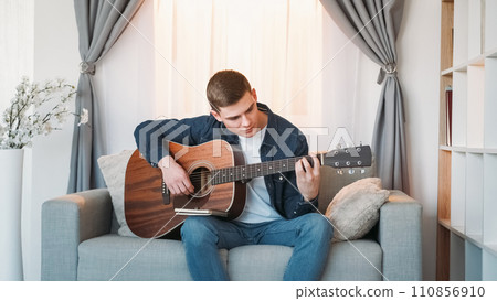 Guitar practicing. Music learning. Motivated inspired man playing melody chords on acoustic string instrument on couch at home in living room. 110856910