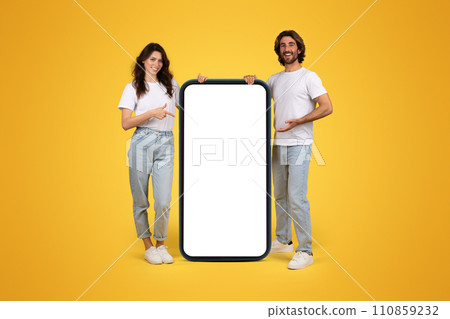 Smiling young european guy and woman in casual point fingers at huge smartphone with empty screen Smiling young european guy and woman in casual point fingers at huge smartphone with empty screen 110859232