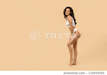 Smiling beautiful woman with sculpted figure posing in lingerie against beige background 110859305