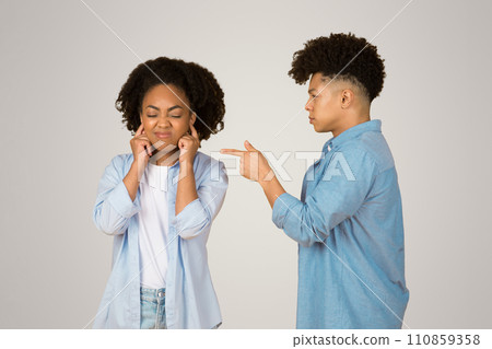 A frustrated young woman covers her ears to ignore a young man pointing at her 110859358