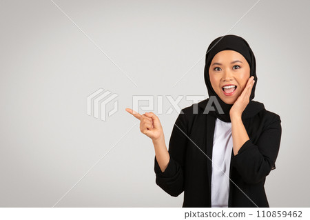 A cheerful young woman in a black hijab and blazer is pointing to the side with one hand while touching her face A cheerful young woman in a black hijab and blazer is pointing to the side with one hand while touching her face 110859462