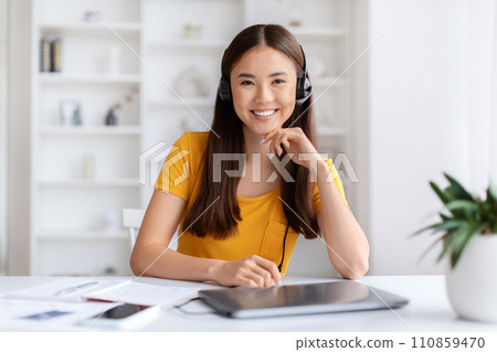 Smiling asian customer service representative woman wearing headset sitting at desk in home office interior, 110859470