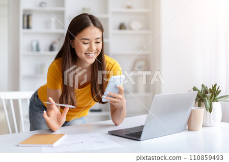 Cheerful asian woman using smartphone while working at desk in home office 110859493