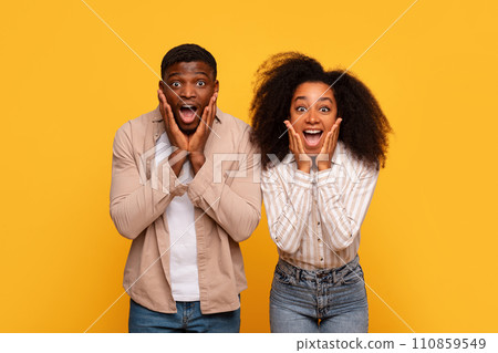 Shocked black couple with hands on cheeks against yellow wall Shocked black couple with hands on cheeks against yellow wall 110859549