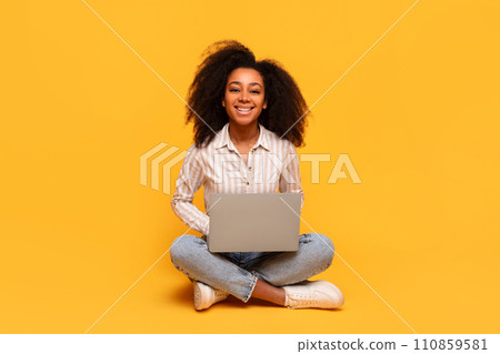 Content black woman with laptop sitting cross-legged on yellow backdrop Content black woman with laptop sitting cross-legged on yellow backdrop 110859581