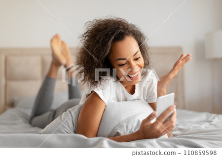 Joyful Black Millennial Lady Communicating Via Cellphone In Bed Indoors 110859598