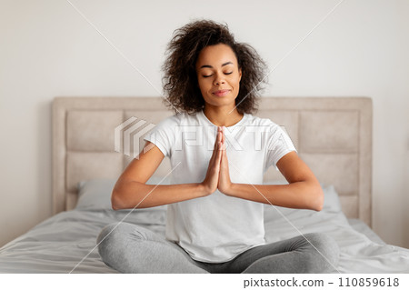 Calm black young woman meditating in lotus position at bedroom Calm black young woman meditating in lotus position at bedroom 110859618