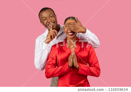 Man with shh gesture, covering woman's eyes, both smiling, pink background 110859634