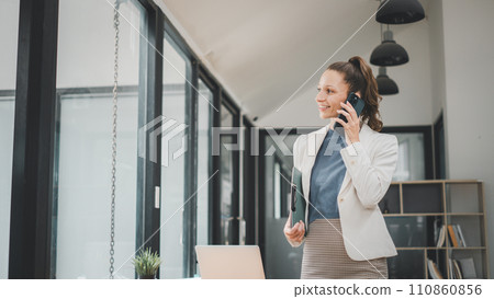 Smiling business woman holding business chart and talking phone standing on modern office background Smiling business woman holding business chart and talking phone standing on modern office background 110860856