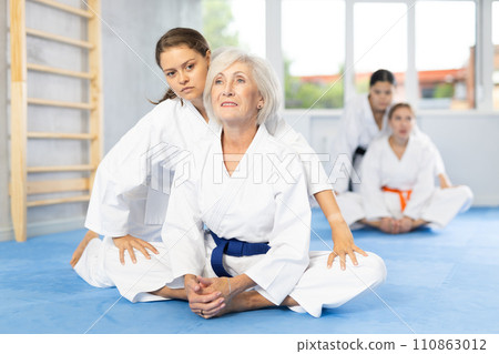Elderly woman in kimono performing butterfly stretch with help of female coach Elderly woman in kimono performing butterfly stretch with help of female coach 110863012