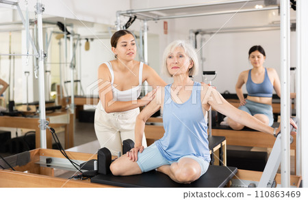 Female pilates instructor helping elderly woman doing exercises on reformer apparatus 110863469