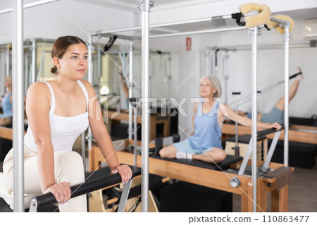 Positive young girl practicing pilates stretching exercises on reformer at gym Positive young girl practicing pilates stretching exercises on reformer at gym 110863477