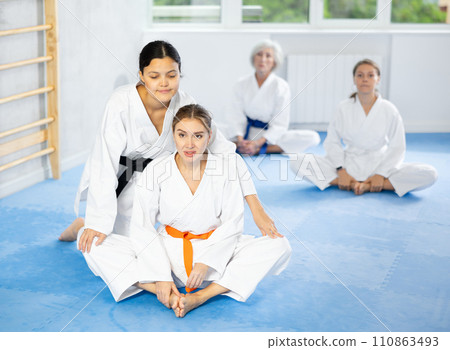 Young woman in kimono stretching in butterfly pose with assistance of trainer Young woman in kimono stretching in butterfly pose with assistance of trainer 110863493
