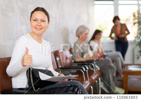 Positive woman showing thumbs up while sitting on a chair in a company office waiting for specialist 110863803