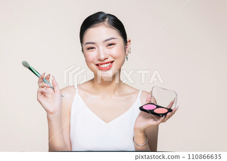 Young asian woman doing makeup with brush while standing on beige background Young asian woman doing makeup with brush while standing on beige background 110866635