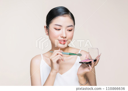 Young asian woman doing makeup with brush while standing on beige background Young asian woman doing makeup with brush while standing on beige background 110866636