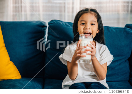 In a cozy apartment a cheerful Asian girl drinks milk from a glass while sitting on the sofa. This heartwarming picture emphasizes daily health care and the joyful innocence of childhood. In a cozy apartment a cheerful Asian girl drinks milk from a glass while sitting on the sofa. This heartwarming picture emphasizes daily health care and the joyful innocence of childhood. 110866848