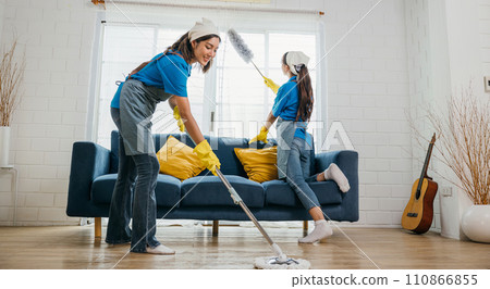 In messy living room two women in uniforms work cleaning employees. With gloves on they tackle dust and dirt on furniture sofas and floors. routine is house care and hygiene. agency small business In messy living room two women in uniforms work cleaning employees. With gloves on they tackle dust and dirt on furniture sofas and floors. routine is house care and hygiene. agency small business 110866855
