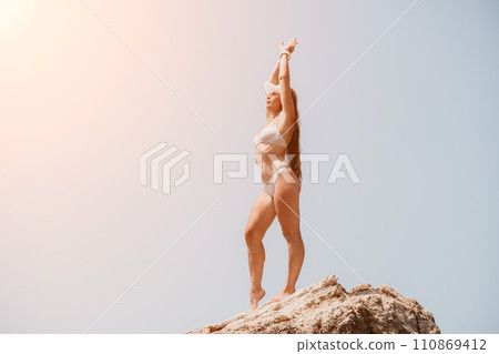 Woman sea yoga. Back view of free calm bliss satisfied woman with long hair standing on top rock with yoga position against of sky by the sea. Healthy lifestyle outdoors in nature, fitness concept. Woman sea yoga. Back view of free calm bliss satisfied woman with long hair standing on top rock with yoga position against of sky by the sea. Healthy lifestyle outdoors in nature, fitness concept. 110869412