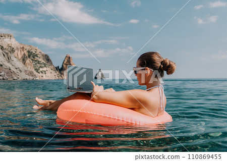 Woman freelancer works on laptop swimming in sea on pink inflatable ring. Happy tourist in sunglasses floating on inflatable donut and working on laptop computer in calm ocean. Remote working anywhere 110869455